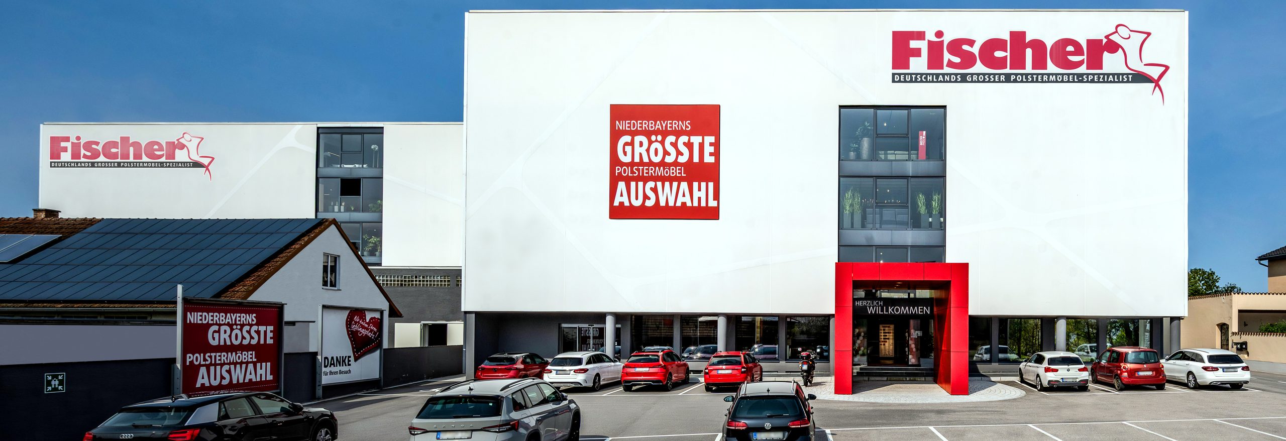 Außenansicht der Filiale Polstermöbel Fischer in Steinach bei Tageslicht, mit rotem Eingangsbereich und geparkten Autos auf dem Kundenparkplatz.