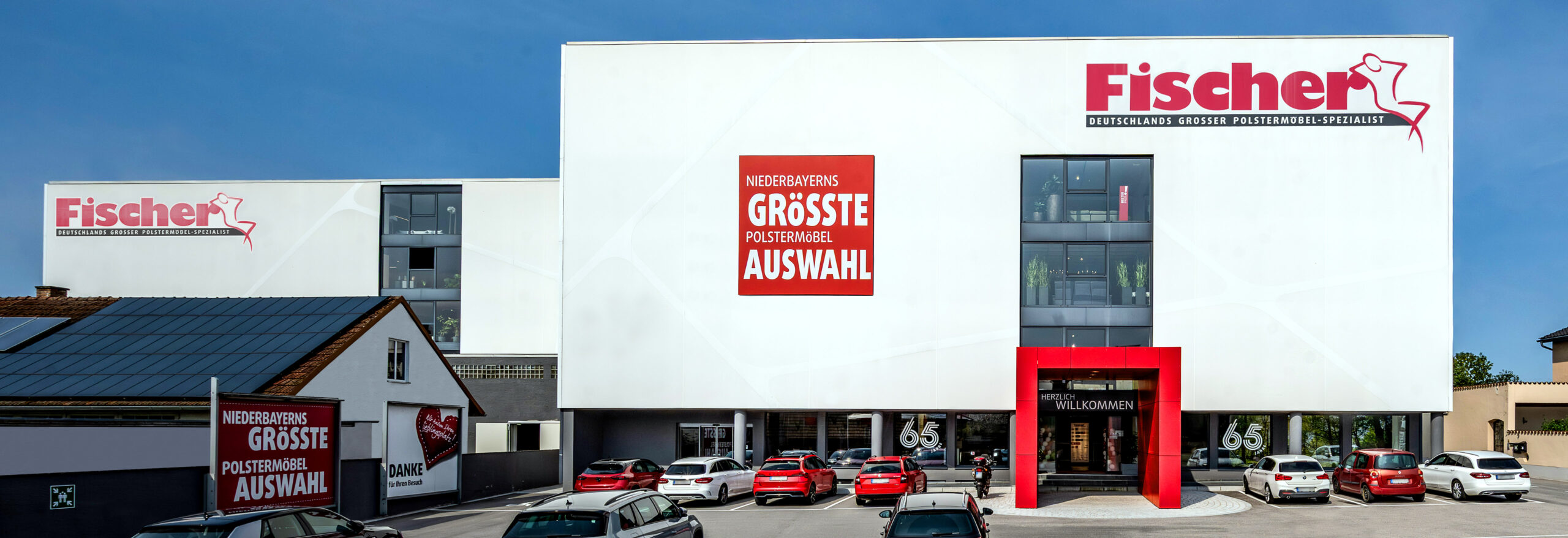 Außenansicht der Filiale Polstermöbel Fischer in Steinach bei Tageslicht, mit rotem Eingangsbereich und geparkten Autos auf dem Kundenparkplatz.