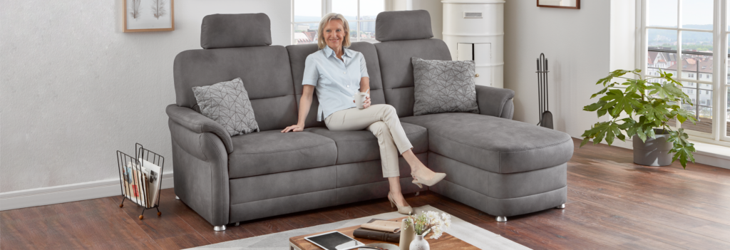 Ältere Frau sitzt auf grauem Sofa mit Kissen.