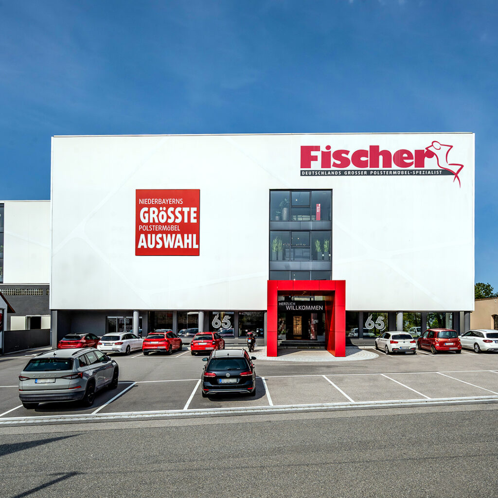 Frontalansicht der Polstermöbel Fischer Filiale in Steinach mit rotem Eingangsbereich, geparkten Autos und großem Fassadenschild „Niederbayerns größte Polstermöbelauswahl“.