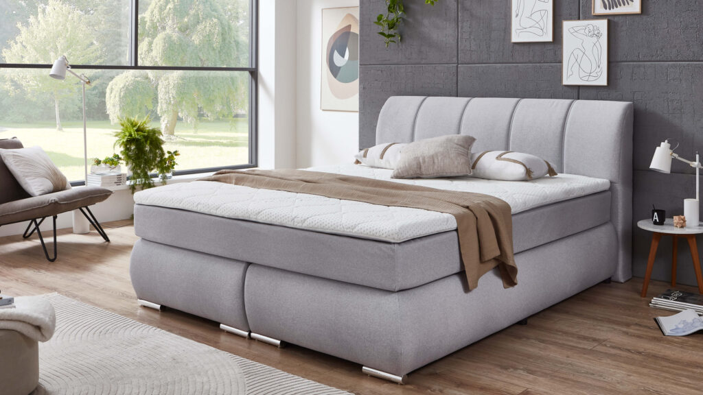 Boxspringbett in gemütlicher Schlafzimmerumgebung, dekoriert mit Kissen und Decke; hellgrauer Stoffbezug und Holzfußboden.