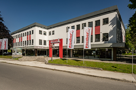 ußenansicht der Polstermöbel Fischer Filiale in Bad Reichenhall – modernes, weißes Gebäude mit roten Akzenten und großem Logo, umgeben von grüner Wiese.