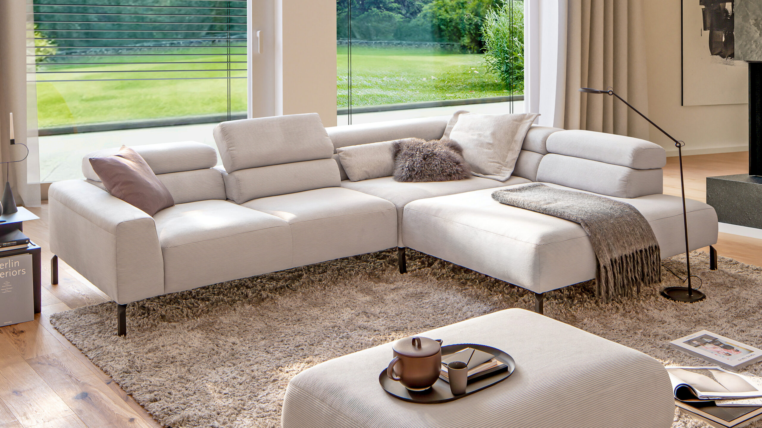 Modernes Ecksofa in hellem Cordstoff mit verstellbaren Kopfstützen in einem großzügigen Wohnzimmer mit Panoramablick ins Grüne.