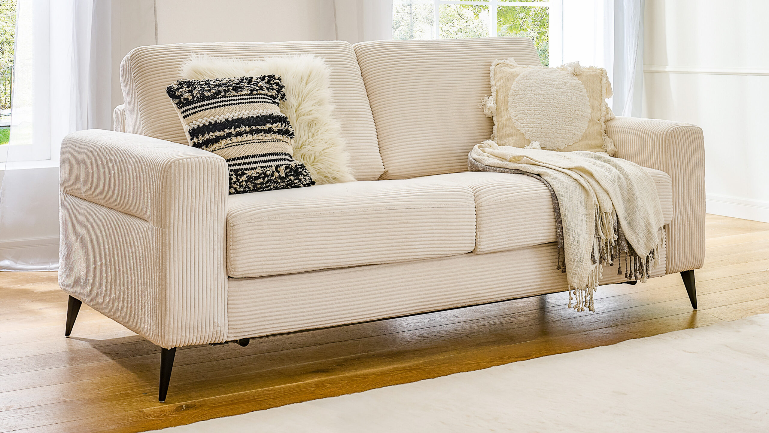 Komfortables Schlafsofa im hellen Corddesign mit geradliniger Form, dekoriert mit Kissen und Plaid in einem lichtdurchfluteten Raum.