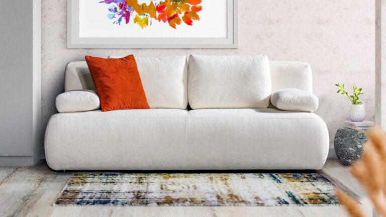 Modernes Schlafsofa in Beige mit rundem Design und orangem Akzentkissen – platzsparendes Sofa für stilvolles Wohnen bei Polstermöbel Fischer.