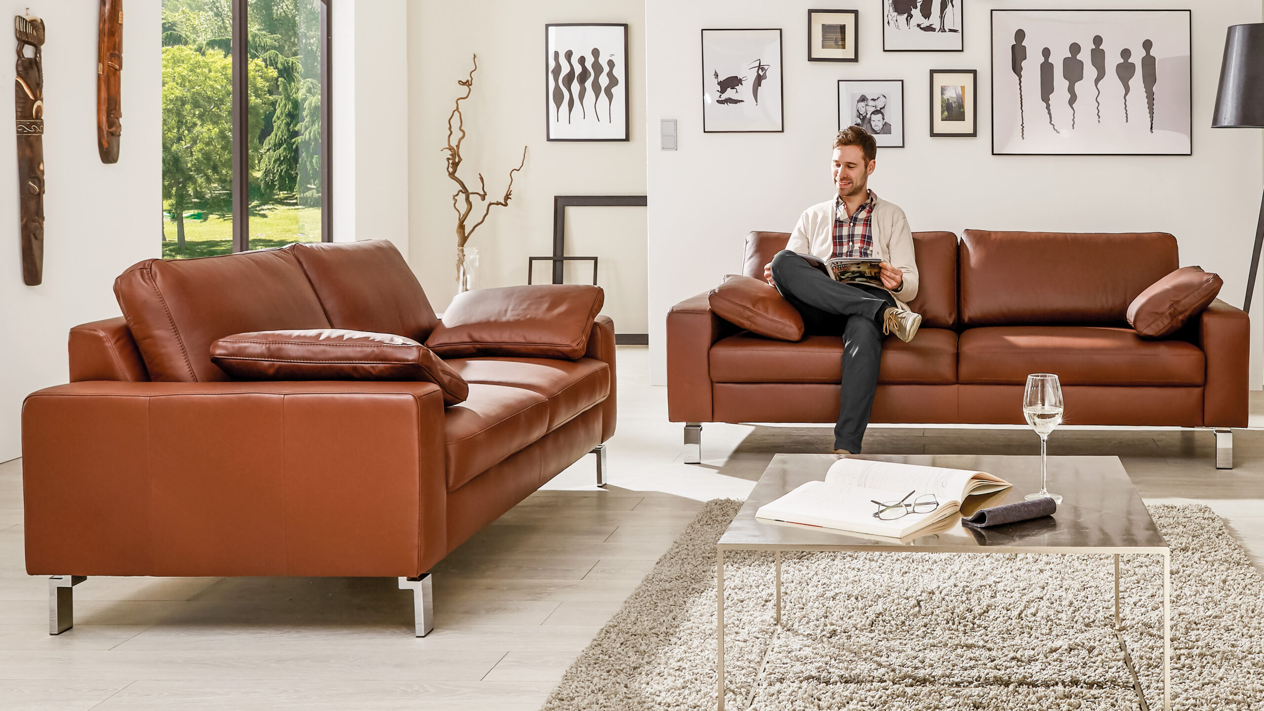 Zwei Einzelsofas aus cognacfarbenem Leder in modernem Wohnzimmerambiente mit Galeriewand und heller Einrichtung, auf einem davon sitzt ein Mann entspannt mit einem Magazin.