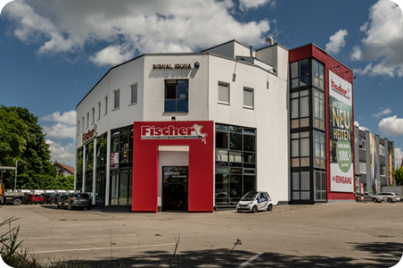 Polstermöbel-Fischer-Filiale in Karlsfeld mit großem Glasbereich, rotem Eingang und angrenzendem Parkplatz bei sonnigem Wetter.