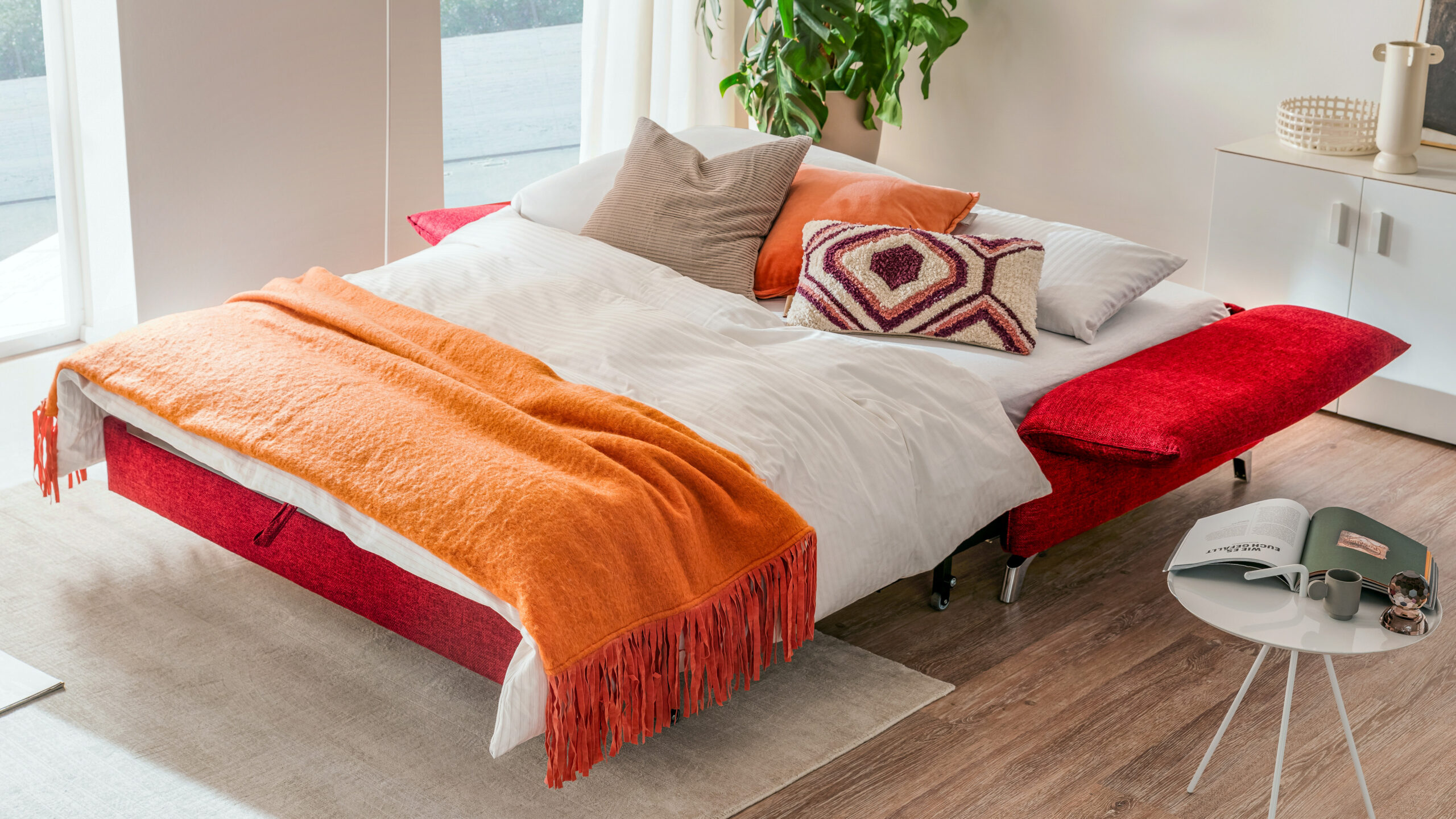Rotes Schlafsofa mit ausgeklappter Schlaffunktion, dekoriert mit Kissen und oranger Wohndecke – gemütliche und farbenfrohe Lösung für Gästeübernachtungen im modernen Wohnraum.