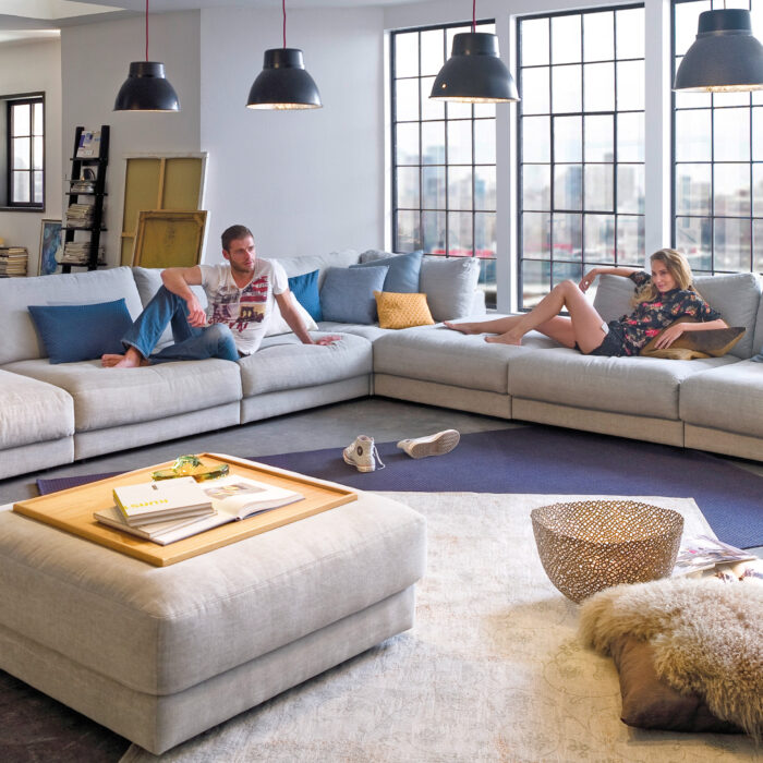 Großzügige Wohnlandschaft in Hellgrau mit vielen Kissen in modernem Loftambiente, zwei Personen sitzen entspannt auf dem Sofa