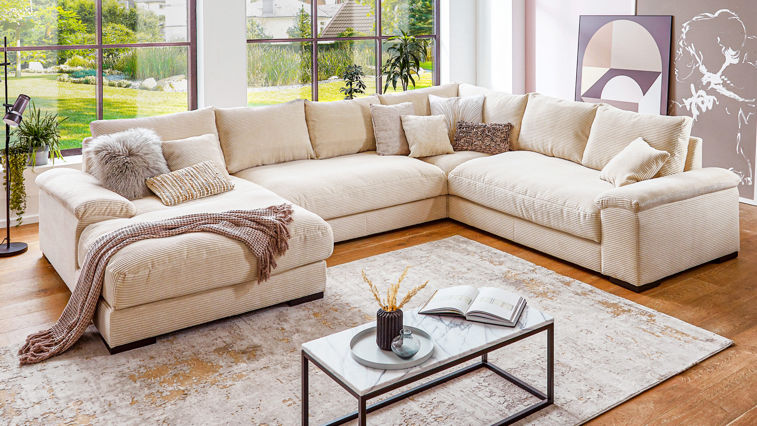 Wohnlandschaft aus beigefarbenem Cordstoff mit vielen Zierkissen und Decken in wohnlicher Einrichtung mit großem Fenster und stilvollem Teppich