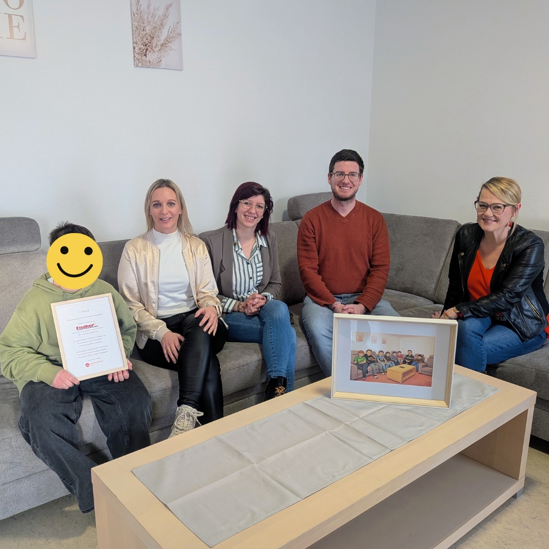 F&uuml;nf Personen sitzen gemeinsam auf einem grauen Sofa in einem hellen Raum. Eine Person h&auml;lt eine gerahmte Urkunde, auf dem Tisch davor steht ein gerahmtes Bild. Die Szene zeigt eine Spendensituation von Polsterm&ouml;bel Fischer an das Antonius Haus Marktl Gruppe Franziskus.